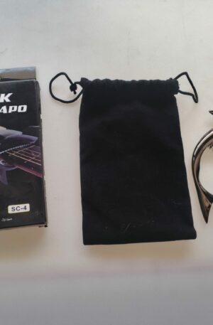 Capodastro de guitarra con diseño de tiburón plateado (Shark Capo SC-4) junto a su caja original y una funda de tela negra sobre fondo claro.